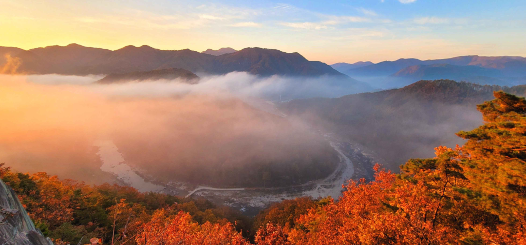 Panoramic view of Korean mountains during autumn with morning fog, red foliage, and sunrise over a valley