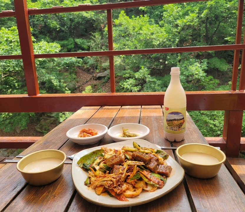 Outdoor Korean mountain restaurant terrace with makgeolli rice wine, and side dishes, overlooking a forested mountainside