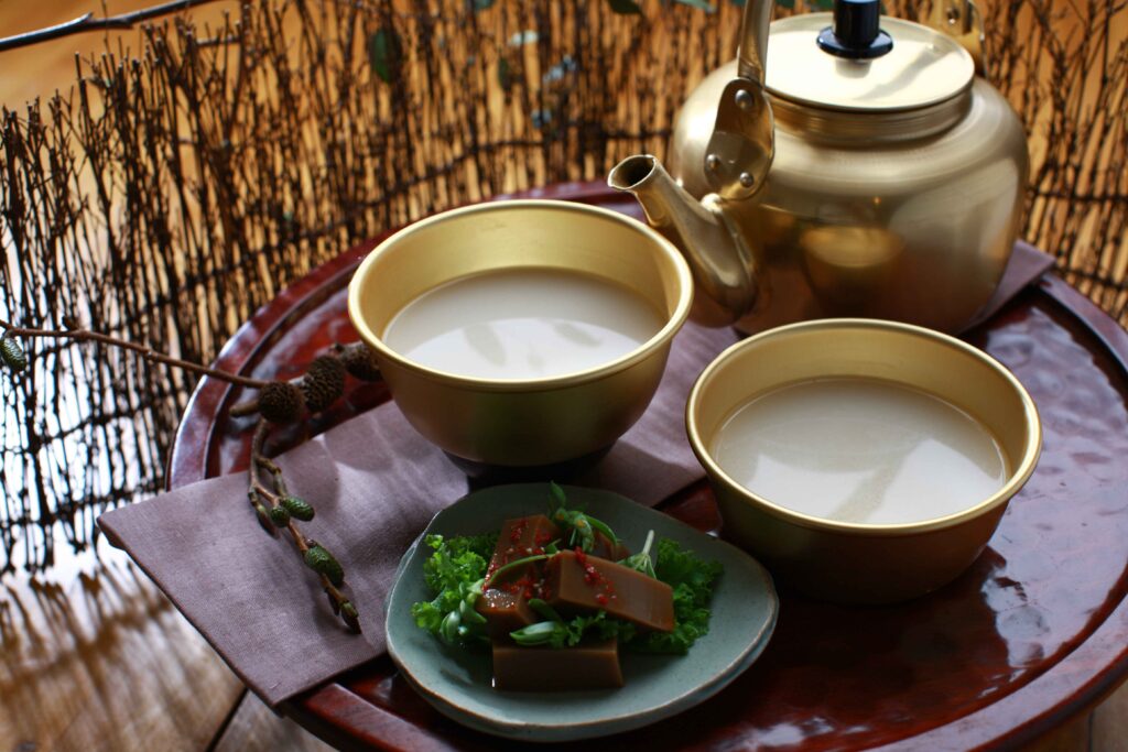 Traditional Korean makgeolli rice wine in brass kettle and **aluminum bowls**, served alongside dotorimuk on a wooden tray