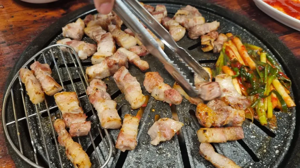 Grilled pork belly pieces being turned with tongs next to seasoned green onion salad