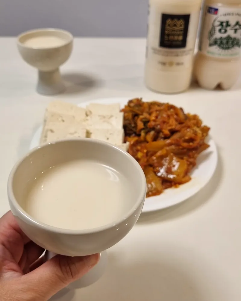 A hand holding a footed ceramic bowl of makgeolli beside a plate of tofu and stir-fried kimchi 