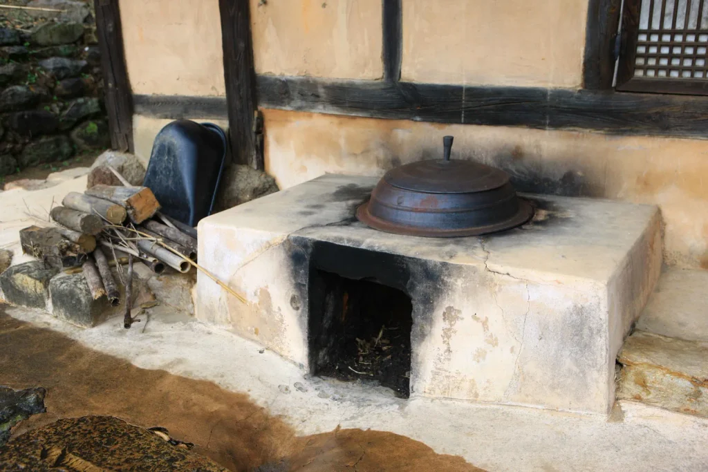 A cast-iron pot resting over the open agungi firebox of a traditional Korean kitchen, with firewood stacked beside it, at Ganggol Village in Boseong