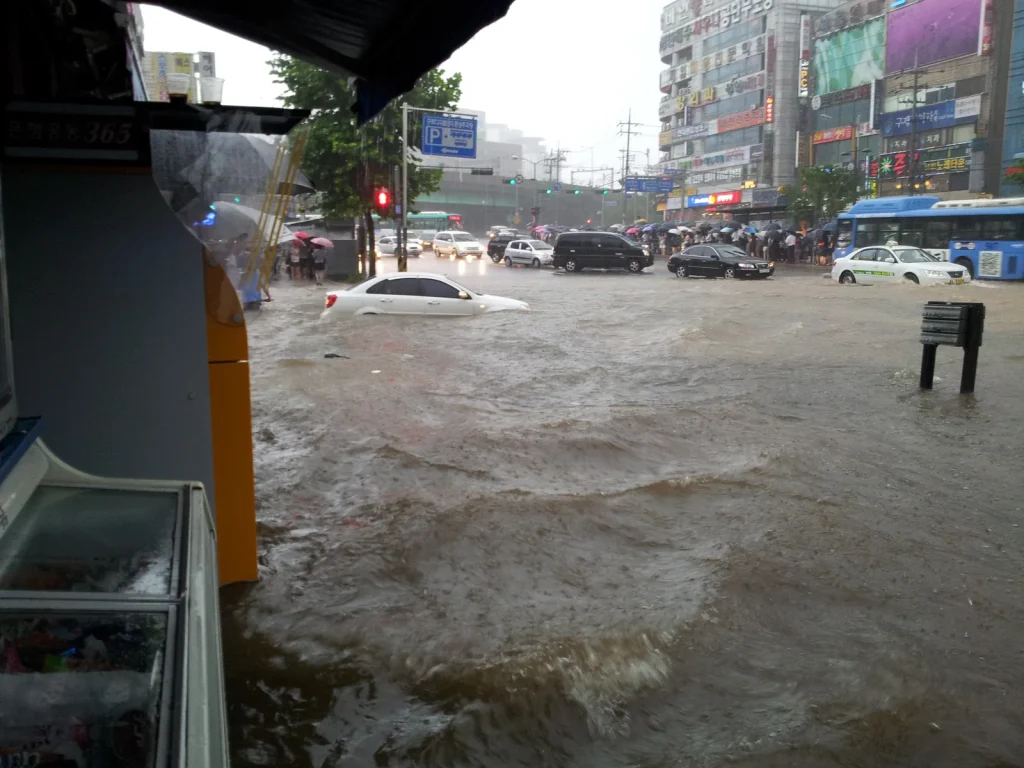 A car half-submerged at a flooded intersection in Guro Digital Complex, Seoul, with buses pushing through brown water and commuters waiting under umbrellas on 27 July 2011